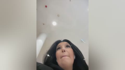 Media: Video of a woman with long black hair, wearing a black dress, looking up with a thoughtful expression, against a white ceiling with scattered lights and a metallic object.