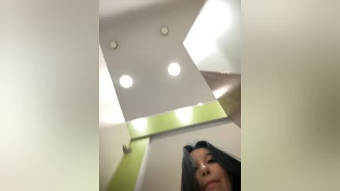 Media: Video of a young woman with long, dark hair, standing in a modern, brightly lit room with white walls, green accents, and recessed ceiling lights.