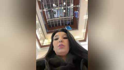 Media: Video of a woman with long black hair, wearing a black coat, standing indoors, looking upward. The background shows a metallic staircase with a wooden railing.