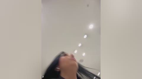 Media: A blurry video of a woman with long, straight black hair, standing in a minimalist, modern white room with recessed lighting. She appears to be looking upwards, wearing a dark top.