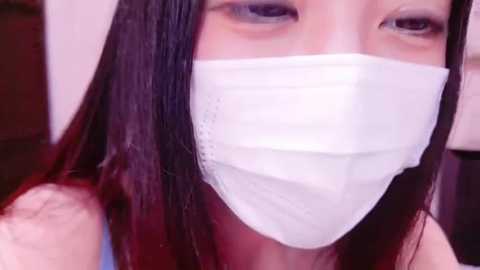 Media: Video of a young woman with long, straight black hair, wearing a white surgical mask, and a blue sleeveless top. Her fair skin and dark eyes are partially visible. Background is blurred, indoor setting.