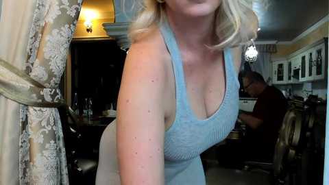 Media: A video of a blonde woman with fair skin, wearing a light blue, sleeveless, ribbed top, leaning forward in a dimly lit, vintage-styled salon with dark wood furniture and chandelier lighting.