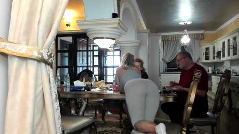 Media: Video of a woman in a tight, gray dress seated on a wooden chair in a lavish, dimly lit dining room with chandeliers, a grand fireplace, and a glass door.