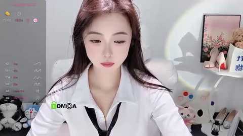 Media: Video of an Asian woman with long, straight black hair, wearing a white lab coat, sitting in a clean, minimalist room with a pink floral painting and a desk with scattered items.