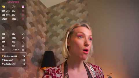 Media: A video of a fair-skinned woman with blonde hair, wearing a patterned blouse, captured on a live stream with a wallpapered background and a black lamp.