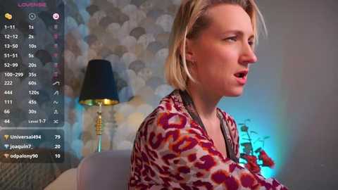 Media: Video of a blonde woman in a leopard-print shirt, sitting in a dimly lit room with a black lamp and floral wallpaper.