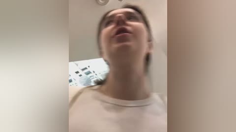 Media: A blurred video of a person with light skin, wearing a beige shirt, looking up with an open mouth, standing in a white room with indistinct, modern decor.