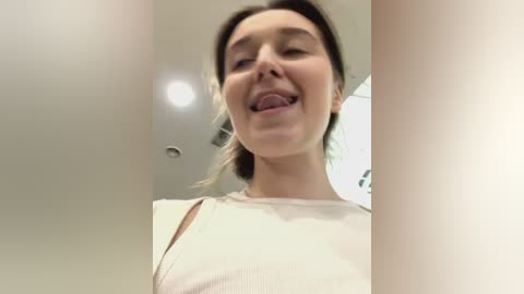 Media: A video of a young woman with light skin and brown hair, smiling and sticking out her tongue, wearing a white ribbed top.