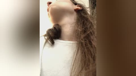 Media: Video of a young woman with long, wavy brown hair, wearing a white ribbed top, arching her back and head back in a sensual pose against a blurred, neutral background.