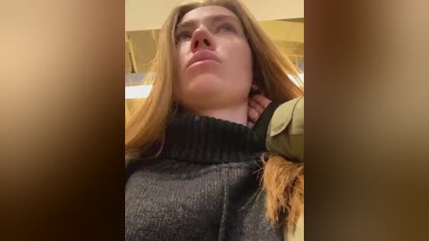 Media: Video of a woman with fair skin and long, straight red hair, wearing a dark grey turtleneck sweater, looking contemplative with a serious expression. Background features blurred, warm-toned textures, possibly wooden surfaces.