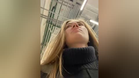 Media: Video of a blonde woman with straight hair, wearing a dark turtleneck sweater, looking up in an indoor setting with metal bars and green paint.