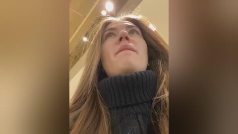 Media: A close-up video of a young woman with long, straight, light brown hair, wearing a dark grey turtleneck sweater, looking upward with a neutral expression. The background features warm-toned, blurry lighting and a ceiling.