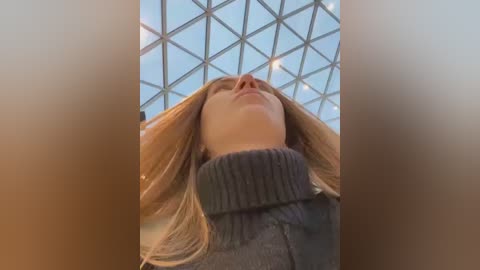 Media: A video of a blonde woman with light skin, wearing a gray turtleneck sweater, standing under a triangular glass ceiling. The image is slightly blurry, focusing on her face and neck.