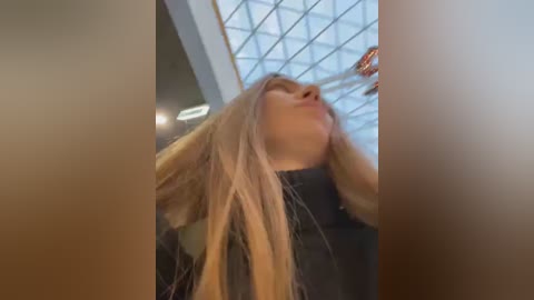 Media: Video of a woman with long, straight, light blonde hair, wearing a black top, reaching up to touch the glass ceiling of a modern, geometrically designed building.