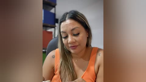 Media: Video of a Latina woman with long, blonde-streaked hair in an orange tank top, sitting in a classroom with blue and red shelves in the background.