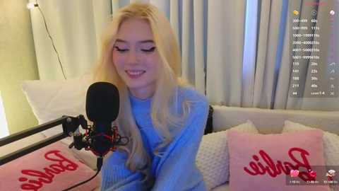 Media: Video of a smiling young woman with long blonde hair, wearing a blue sweater, singing into a microphone, surrounded by pink pillows and white curtains.