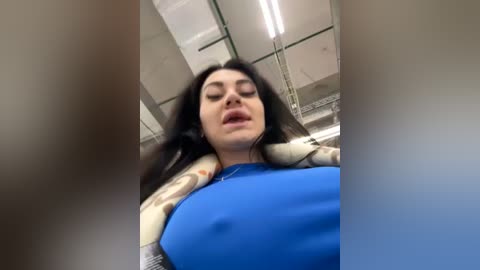 Media: A video of a woman with long dark hair, wearing a blue top and a plaid jacket, taken from a low angle, showing her cleavage and a blurred background of a modern office space with fluorescent lights.