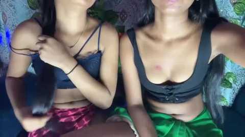 Media: Video of two young women with medium skin tones, wearing black crop tops and red plaid skirts, sitting closely together, showing a slight nip slip.