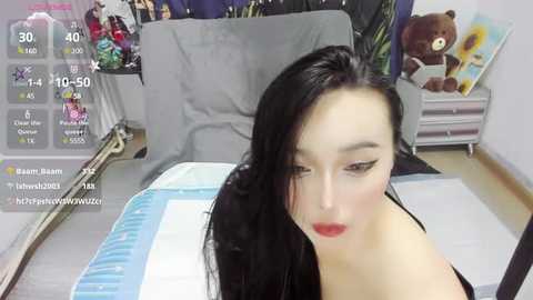 Media: A video of a young Asian woman with long black hair and red lipstick, sitting on a bed in a bedroom with plush toys and a TV showing game statistics.