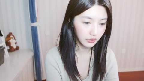 Media: A video of a young Asian woman with long black hair and fair skin, wearing a light grey top, seated indoors. Background shows a beige wall, a blue column, and a stuffed toy bear.