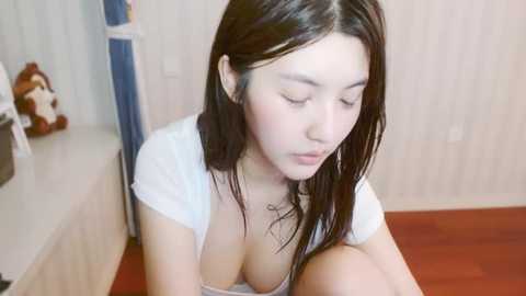 Media: A video of an Asian woman with long black hair, fair skin, and medium-sized breasts, wearing a white top, sitting on a wooden floor. The background features a stuffed bear and a blue curtain.