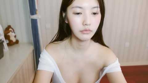 Media: A video of an Asian woman with long dark hair, fair skin, and small breasts, wearing a white off-shoulder top, sitting in a simple room with light walls and a stuffed animal in the background.