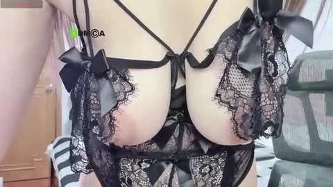 Media: Video of a woman in black lace lingerie with sheer fabric, revealing her large breasts, with a green label \"GA\" on her left shoulder. Background includes a wooden desk and green office chair.