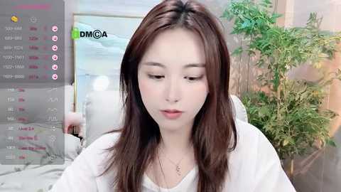 Media: Video of an Asian woman with long, straight brown hair, wearing a white top, sitting in a cozy room with green plants, a bed, and a mirror in the background.