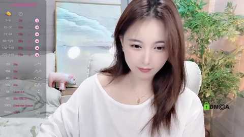 Media: Video of a young Asian woman with long brown hair, fair skin, wearing a white off-shoulder sweater, sitting indoors. Background includes a TV showing a beach scene, a green potted plant, and a digital clock.