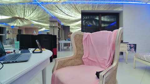 Media: Video of a modern, minimalist office with white walls, blue LED strip lighting, and a pink-wrapped armchair, featuring a desk cluttered with a laptop, papers, and a cup.