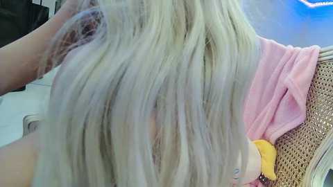 Media: Video of a blonde woman with long, straight hair, sitting in a chair, undergoing hair dye application. Her hair is wet, and she's wearing a pink shirt. Background shows a wicker chair and a blue light.