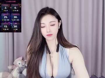 Media: Video of a young Asian woman with long, straight dark hair, fair skin, and large breasts, wearing a low-cut light blue halter top. She sits in a room with a plush toy and gray curtains in the background.