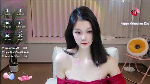 Media: A video of a young Asian woman with long black hair, fair skin, and red lipstick, wearing a red off-shoulder top, sitting in a modern room with a pink floor lamp, a white chair, and a window with blinds.