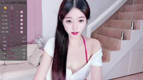 Media: Video of an East Asian woman with long black hair, wearing a white off-shoulder top, revealing a pink bra, posing indoors near a staircase.