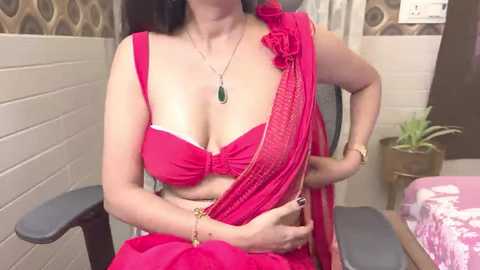 Media: Video of a South Asian woman with medium skin tone wearing a vibrant pink sari, revealing cleavage in a red bra, sitting on a gray chair in a tiled room.