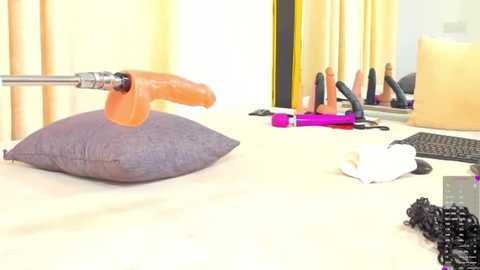 Media: Video of a modern office desk with a gray cushion, an orange dildo, pink vibrator, and black keyboard. The background features beige curtains and a computer monitor.