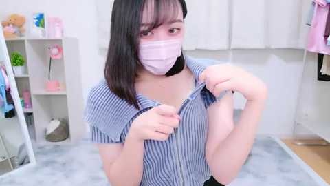 Media: A video of an Asian woman with light skin, wearing a blue and white striped shirt, a pink face mask, and a black choker, adjusting her shirt in a bright, cluttered room.