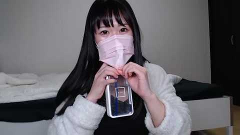 Media: Video of an Asian woman with long black hair and a pink surgical mask, wearing a white fluffy robe, holding a clear plastic container, in a simple, white-walled room.