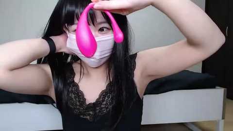 Media: Video of an East Asian woman with long black hair, wearing a pink face mask, black lace top, and pink dildo, posing seductively in a bedroom with white furniture.