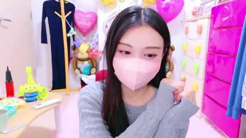 Media: Video of an Asian woman with long black hair, wearing a pink mask and gray sweater, holding a pink object, in a brightly lit, colorful, and organized room filled with toys and clothes.