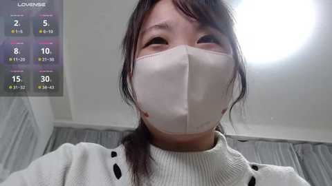 Media: A video of an East Asian woman with long dark hair, wearing a white face mask and a cream sweater, taken indoors with a ceiling light.