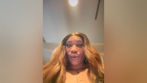 Media: Video of a young Black woman with long, wavy brown hair and light brown skin, wearing a neutral-colored top. She has a playful expression, sticking out her tongue. The background is a dimly lit, beige room with a ceiling light.