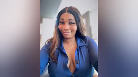 Media: Video of a smiling, dark-skinned woman with long, wavy hair, wearing a low-cut, blue blouse, revealing cleavage, in a softly blurred, indoor setting.