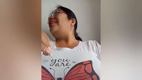 Media: Video of a smiling woman with glasses, medium skin tone, dark hair, wearing a white T-shirt with a red butterfly graphic and \"you dare\" text, against a plain background.