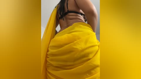 Media: A video of a woman in a bright yellow saree, standing with her back to the camera, showcasing her curvy hips and black bra strap. The background is blurred, focusing on her attire.