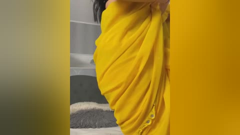 Media: Video of a person in a bright yellow sari, partially draped, standing beside a grey and white bed with a fluffy grey pillow.