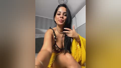 Media: Video of a Latina woman with long black hair, wearing a yellow robe, black bra, and necklace, posing provocatively in a modern bathroom with tiled walls.