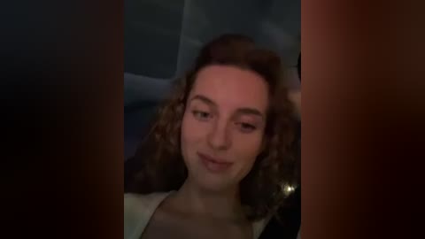 Media: Video of a smiling woman with curly brown hair and light skin, wearing a white top, seen from a dimly lit car interior.