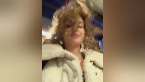 Media: A blurry video of a woman with curly brown hair, wearing a white fur-trimmed coat, smiling, with a blurred background.