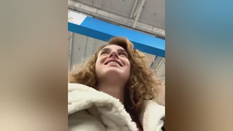 Media: A video captures a young woman with curly blonde hair, smiling broadly, wearing a white fluffy robe. The background features a gymnasium with blue walls and white ceiling. The image is taken from a low angle, emphasizing her happiness.